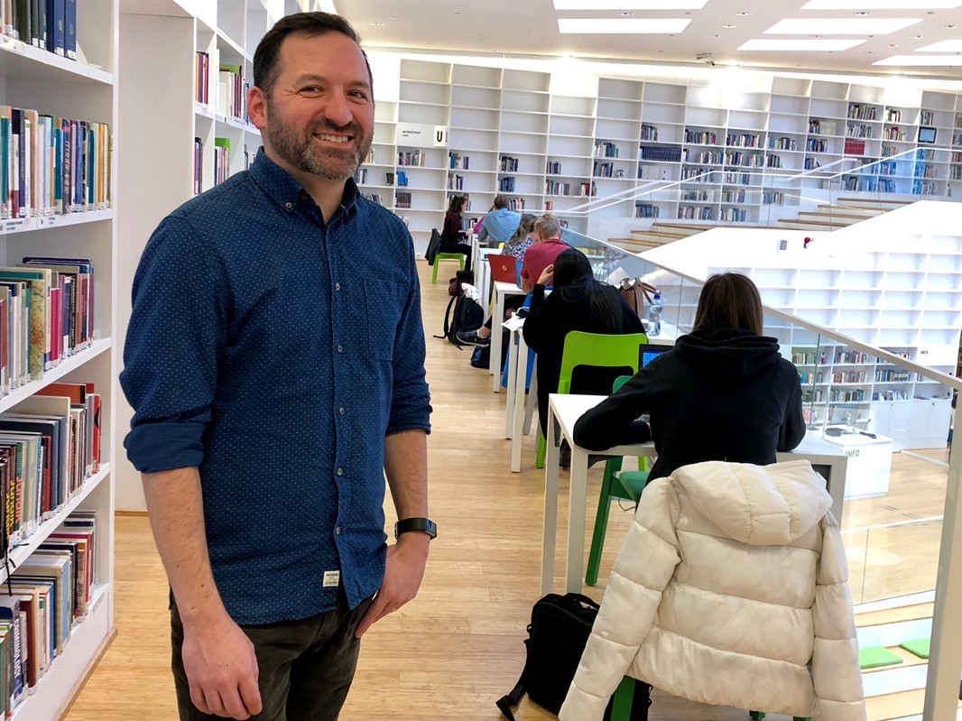 Michael Oppenheimer i blå jeansskjorta ler mot kameran i högskolans bibliotek.
