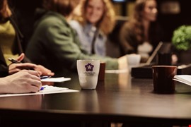 Mugg med Högskolan Dalarna logotyp bord inomhus personer i bakgrunden