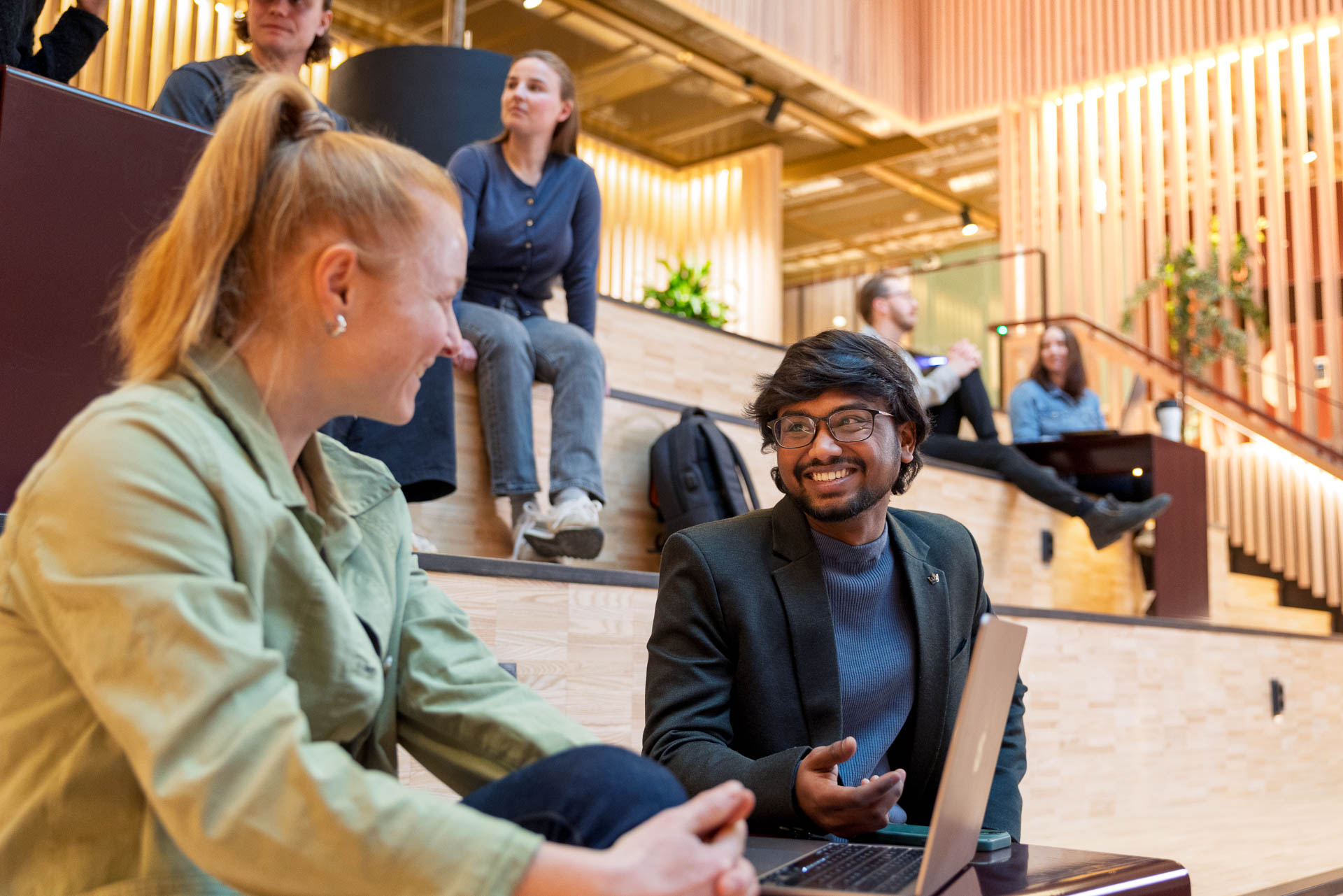 Två studenter samtalar vid en dator i Kunskapstrappan på Campus Borlänge. I bakgrunden syns fler studenter i trappan.