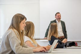 En lärare står framme vid en whiteboard i ett klassrum. Tre studenter sitter framför.