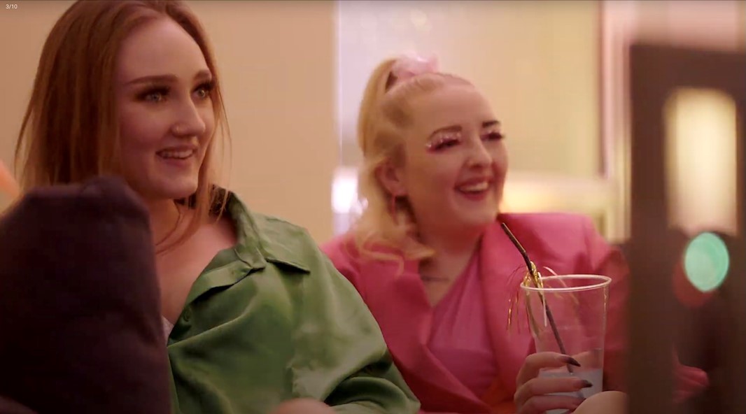 Två unga leende kvinnor sitter i färglada kläder i en soffa på en fest.