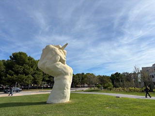 Large statue of a white hand with a pen pointing to the sky on a lawn with people walking by on paths