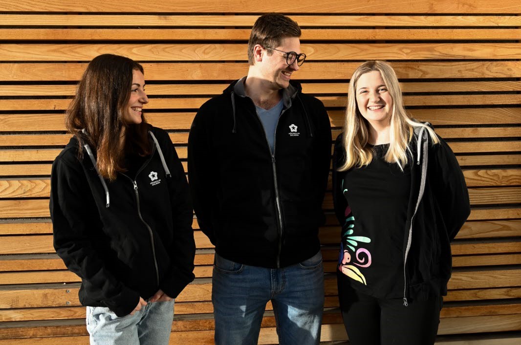 Tre studenter som står mot en trävägg och ler.