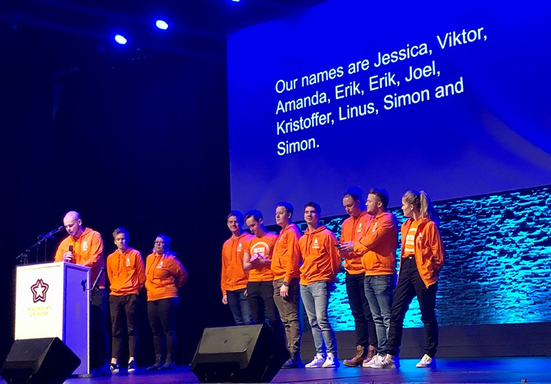 Students in orange shirts on the stage introducing themselves to the audience.