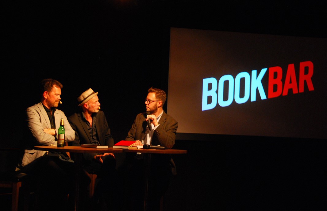 The two authors and the chief editor on the stage with a projected image showing the words BOOKBAR behind them.