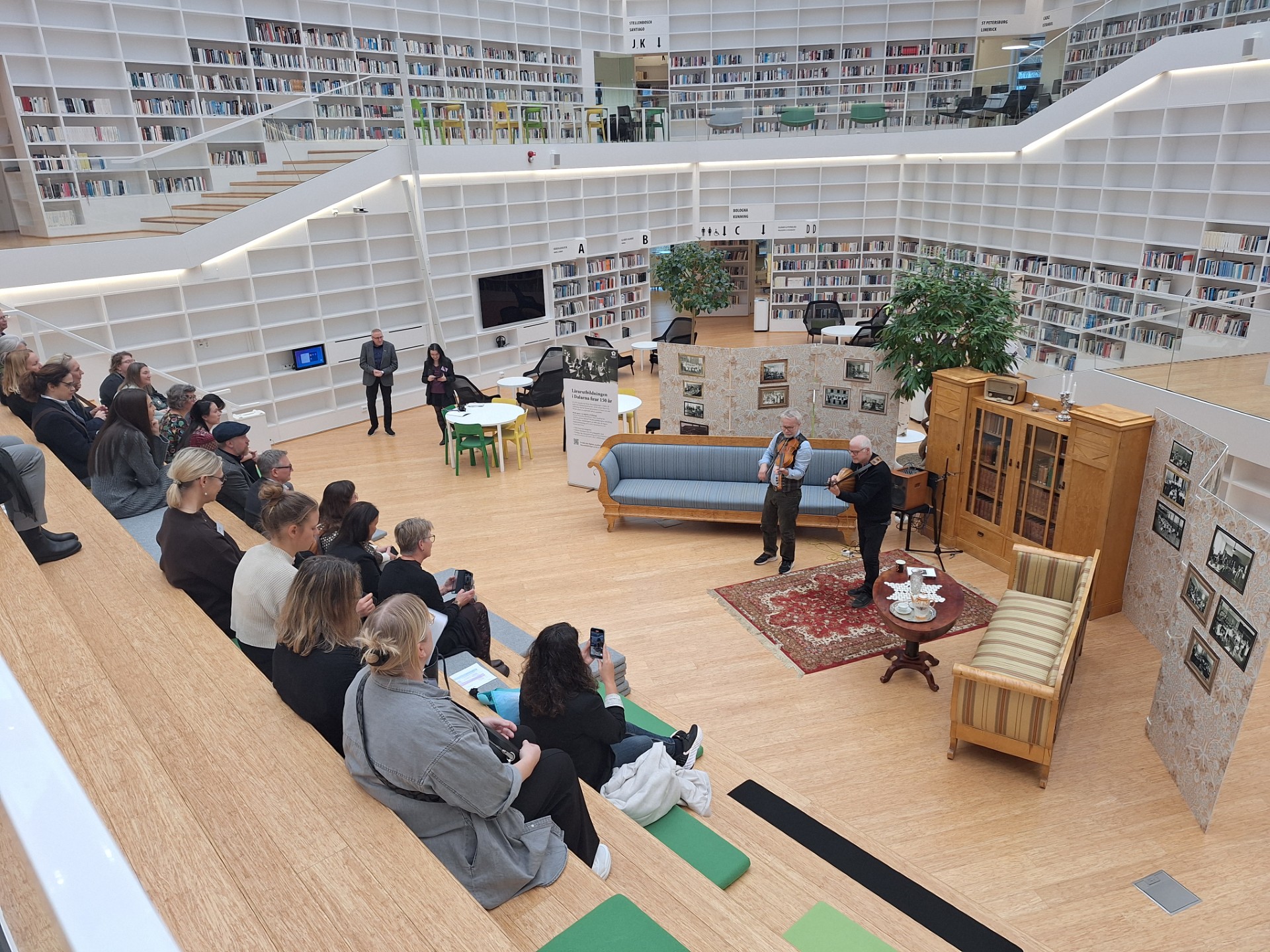 En grupp besökare sitter i biblioteket där det står några musikanter på scenen med gamla möbler