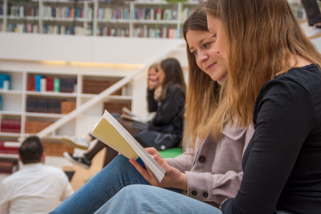 Fyra studenter sitter två och två. Ena paret läser bok tillsammans medan det andra paret samtalar med en lärare.