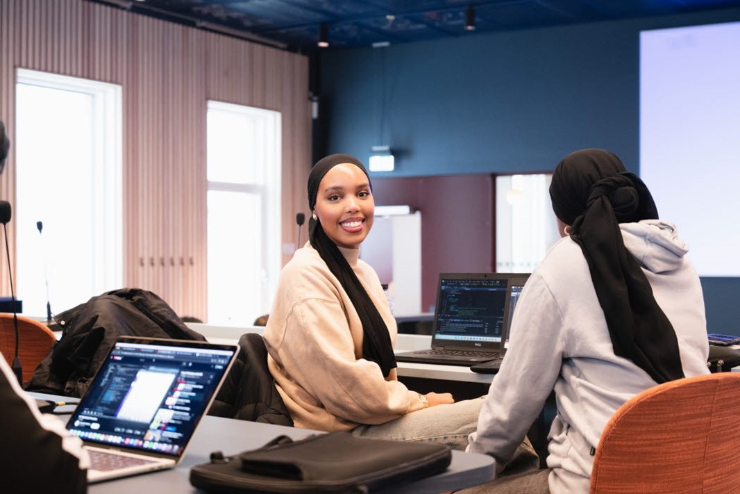 Studenter i ett klassrum som studerar vid sina datorer. En av studenterna kollar rakt in i kameran. 