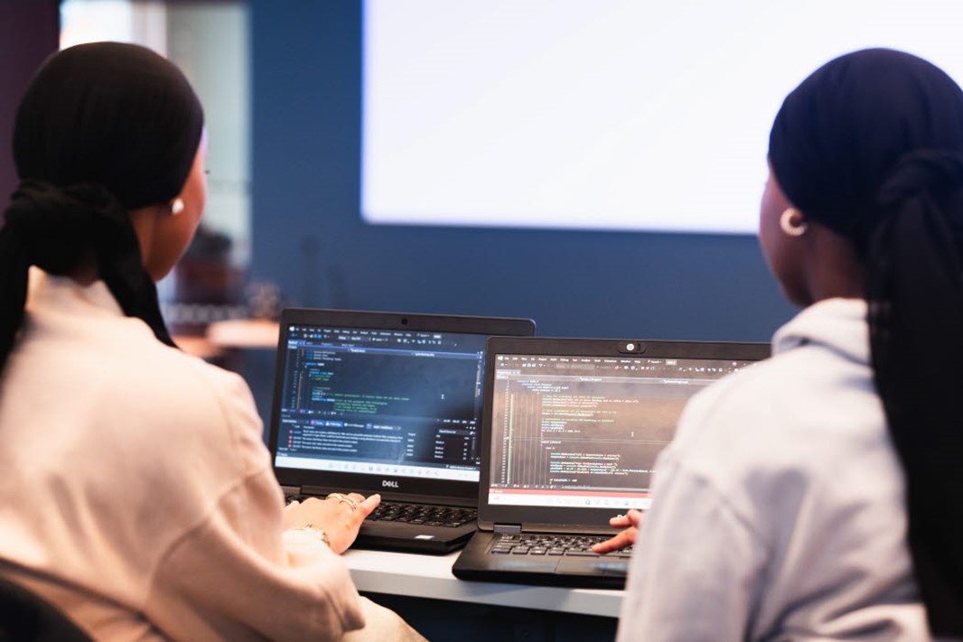 Två studenter som sitter med ryggen vänd mot kameran, båda håller på att programmera på sina datorer. 