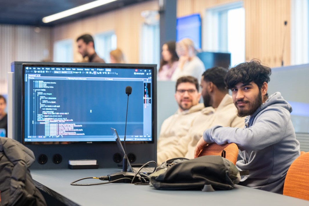 Studenter som sitter i ett klassrum. Fokus ligger på en student som sitter med sin laptop och kollar rakt in i kameran