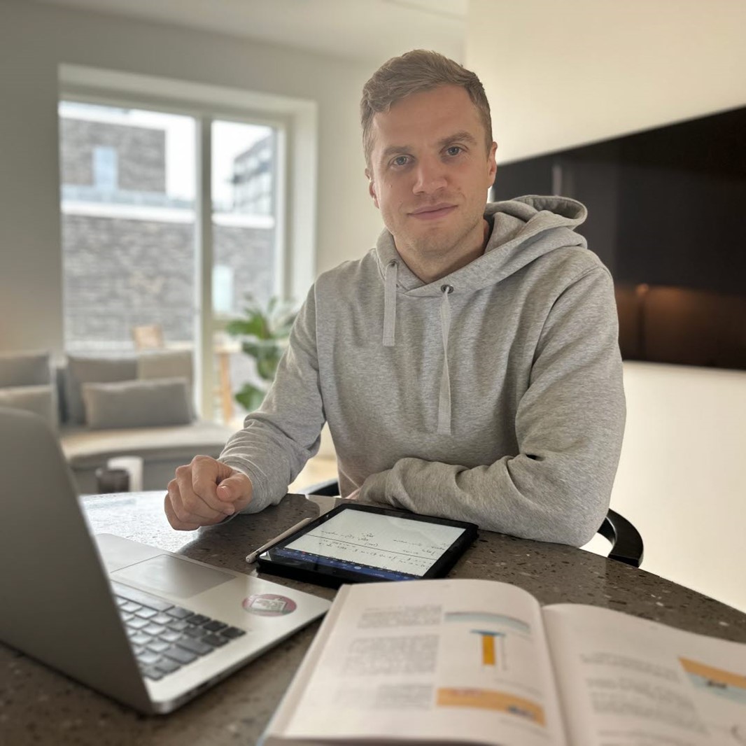 Kille som sitter vid ett bord hemma framför en dator och en bok liggandes på bordet.